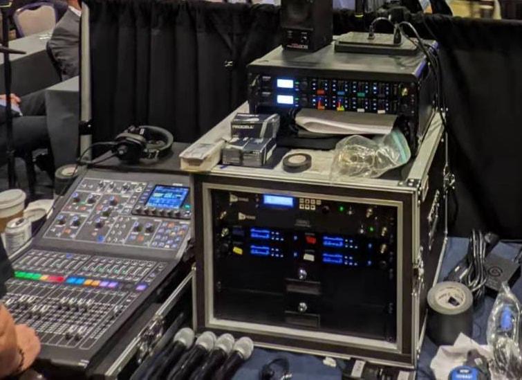 A digital mixing console, a rack of wireless mic receivers and handheld mics at front of house