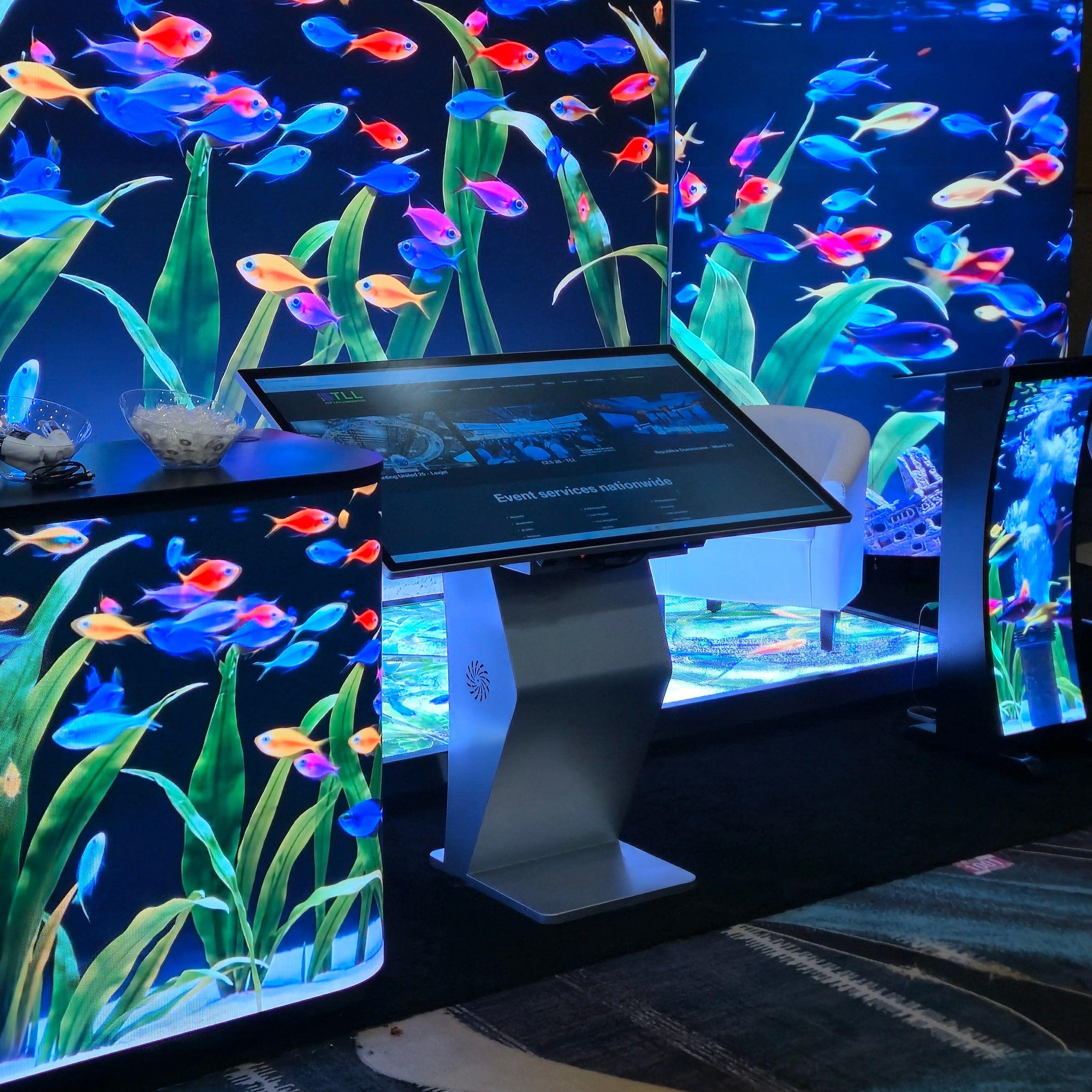 A tilt-top touchscreen kiosk in front of LED walls and an LED counter, all playing a bright aquarium scene