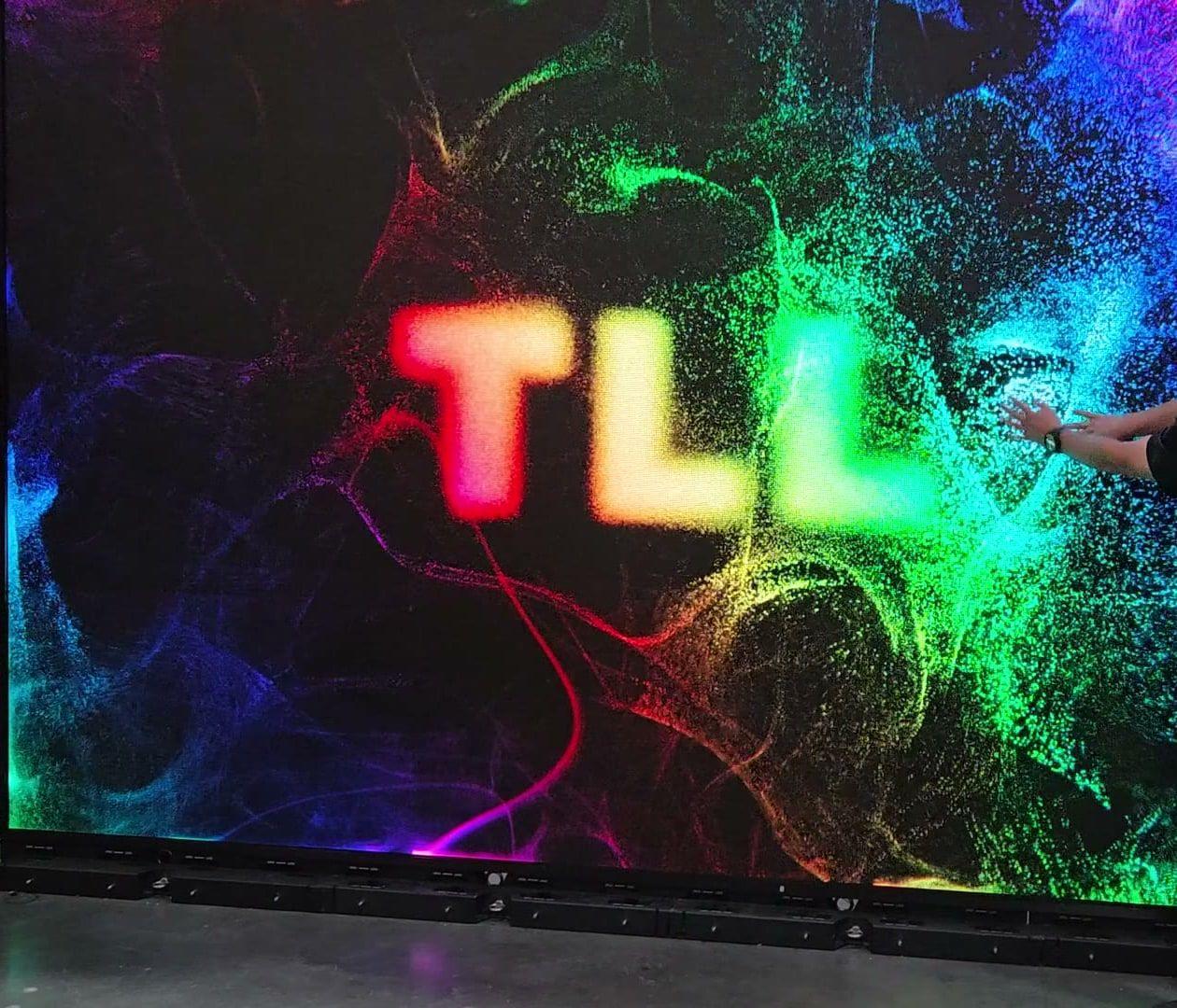 Hands touching a large LED wall as colored particles swirl around the letters TLL